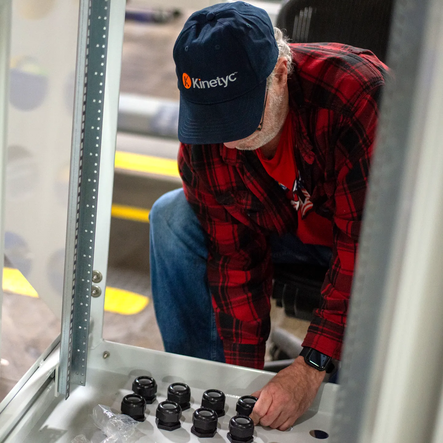 A worker in a red plaid shirt and a Kinetyc cap assembling a customer's product, carefully installing components inside a metal enclosure.