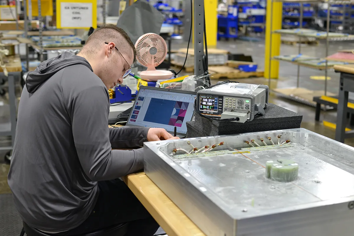 A technician working at a manufacturing workstation, using a laptop and testing equipment to inspect an aluminum component.