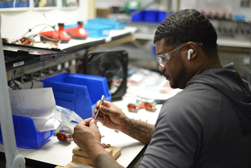 A technician assembling a component at a workstation, using precision tools while wearing safety glasses and earbuds.