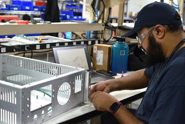 A Kinetyc employee assembling an electronics enclosure at a workstation, following design specifications on a computer screen.