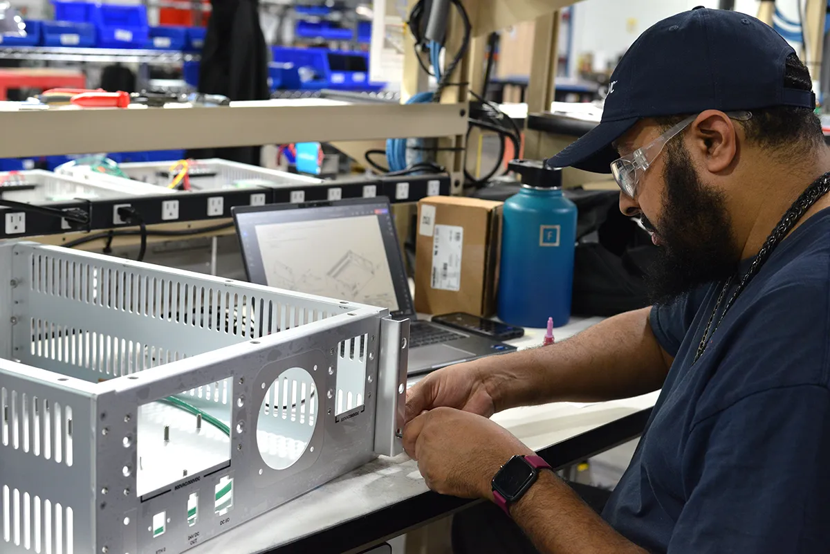 A Kinetyc employee assembling an electronics enclosure at a workstation, following design specifications on a computer screen.