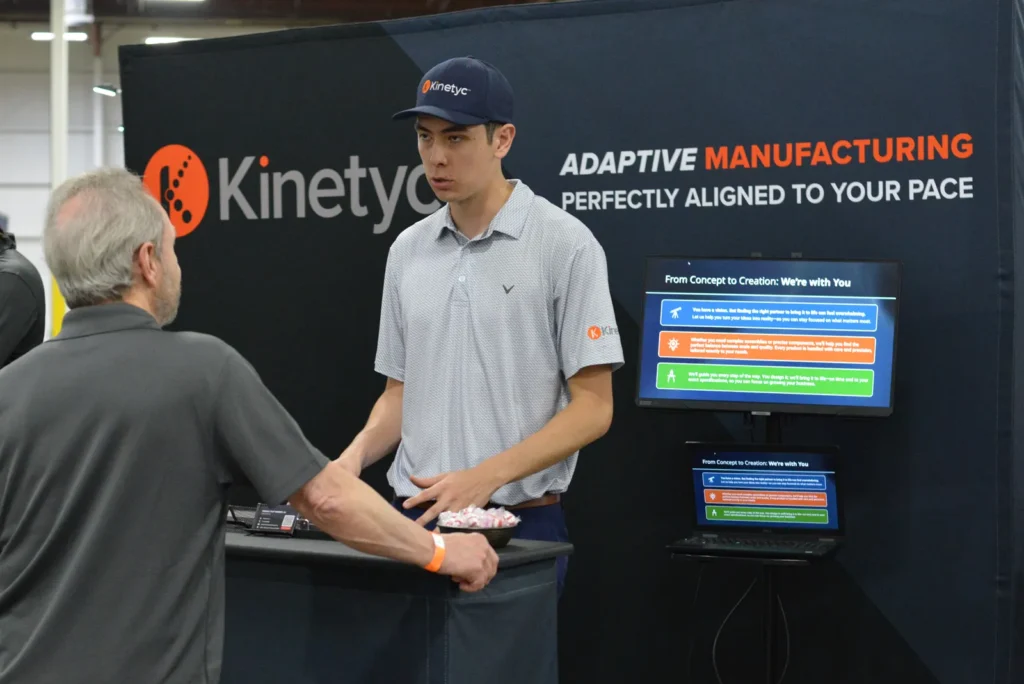 A Kinetyc team member speaks with a visitor at a trade show booth showcasing adaptive manufacturing solutions