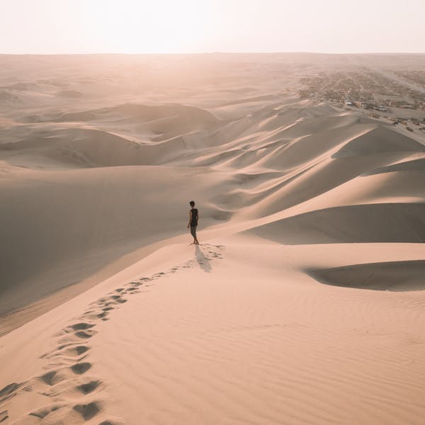 A lone traveler walks across the vast sand dunes of Ica, Peru, under the warm glow of a sunset.