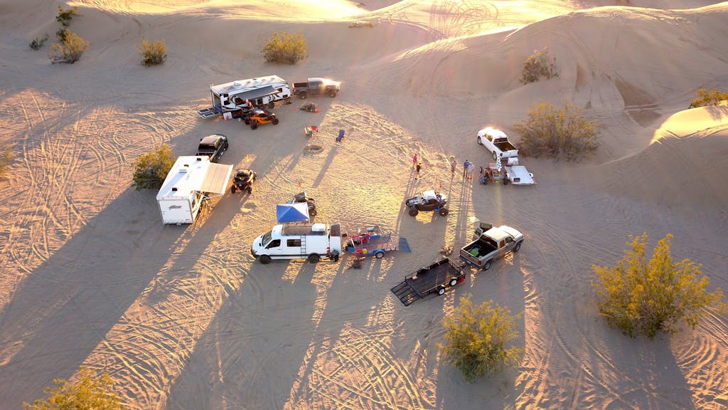 High-angle drone shot of a desert campsite with vehicles and trailers at sunset.